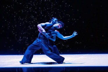 Dance Me – Music by Leonard Cohen Two dancers move closely together on a dark blue stage with falling particles.