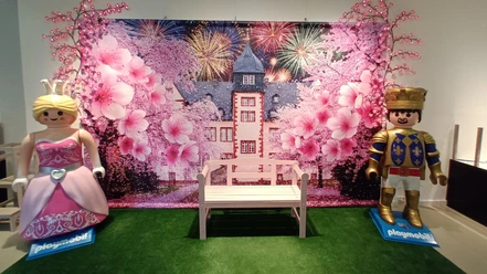 Playmobilausstellung_Salder_HD_16zu9.jpg Playmobil-Prinzessin und Ritter neben einer Bank vor Schlosskulisse mit Blüten.