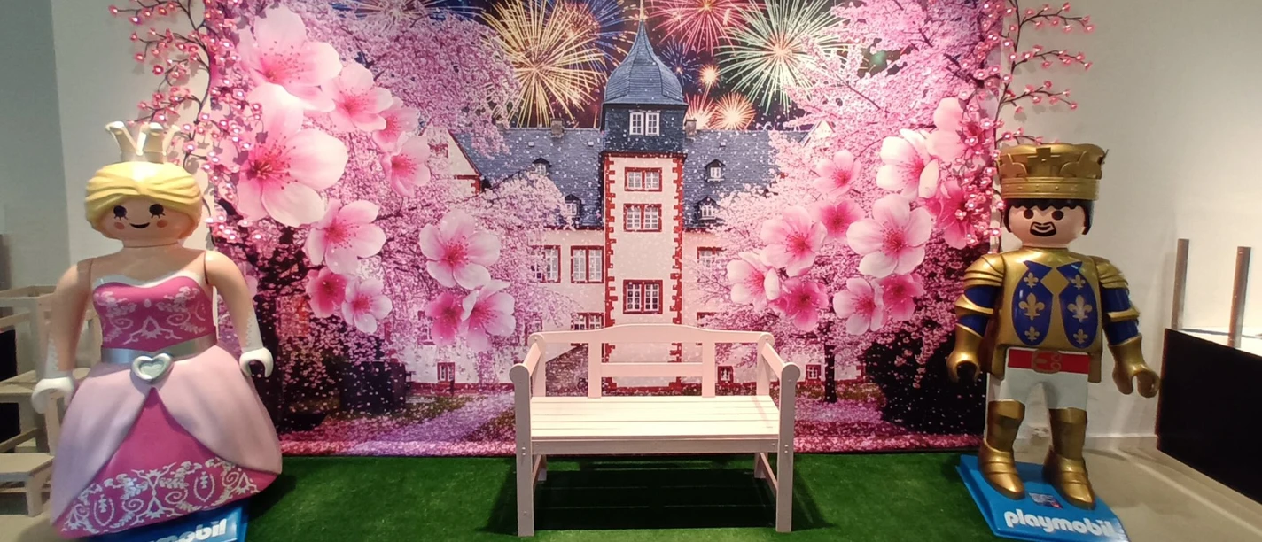 Playmobilausstellung_Salder_HD_16zu9.jpg Playmobil-Prinzessin und Ritter neben einer Bank vor Schlosskulisse mit Blüten.