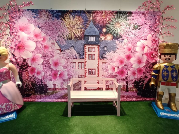 Playmobilausstellung_Salder_HD_16zu9.jpg Playmobil-Prinzessin und Ritter neben einer Bank vor Schlosskulisse mit Blüten.
