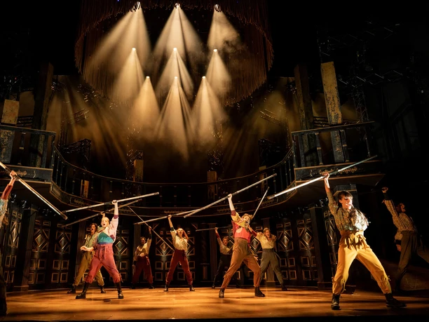 ROMEO & JULIA – LIEBE IST ALLES Tänzerensemble hebt Stangen im warmen Scheinwerferlicht einer kraftvollen Bühneninszenierung.