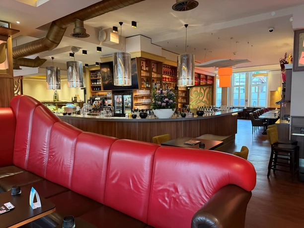 Cup&Cino 2026 Rotes Ledersofa vor einer runden Holztheke im modernen Innenraum eines Cafés.
