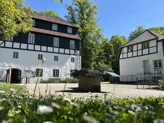 LVR-Industriemuseum Papiermühle Alte Dombach Zwei Fachwerkgebäude mit Innenhof und steinernem Bottich vor blauem Himmel und grüner Kulisse.