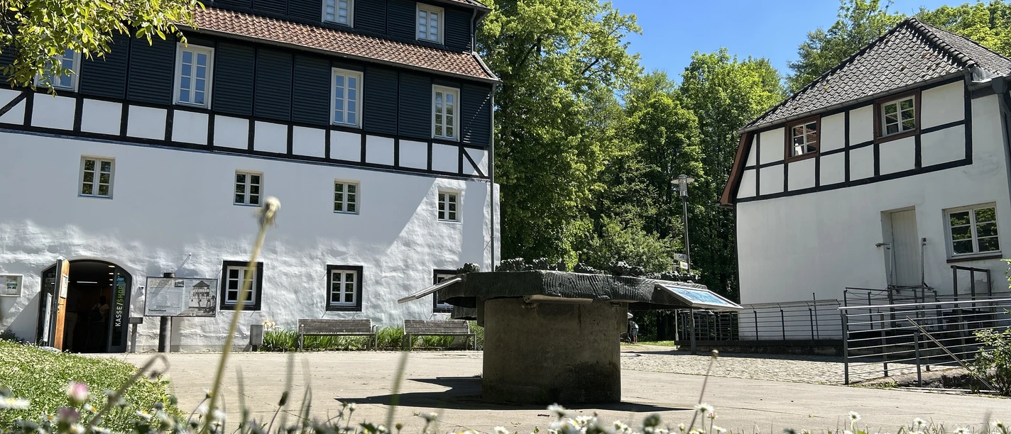 LVR-Industriemuseum Papiermühle Alte Dombach Zwei Fachwerkgebäude mit Innenhof und steinernem Bottich vor blauem Himmel und grüner Kulisse.