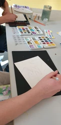 Auf dem Bild sind Menschen dargestellt, die mit einem Pinsel Aquarellbilder auf weißes Papier malen.