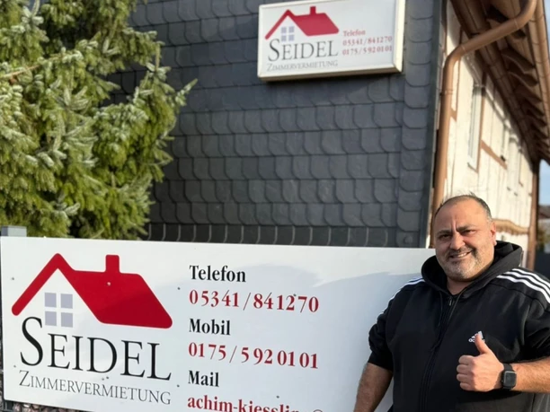 Lächelnder Mann mit Daumen nach oben vor dem Schild der Zimmervermietung Seidel.