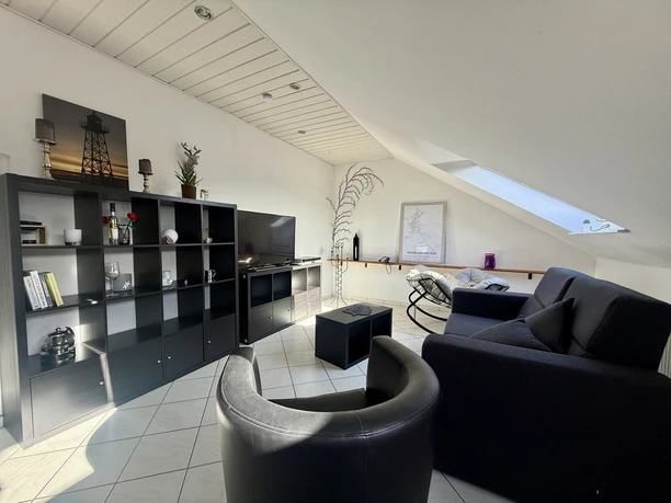 Helles Wohnzimmer mit Dachschräge, schwarzem Sofa, Sessel, Regalwand und Fernseher.