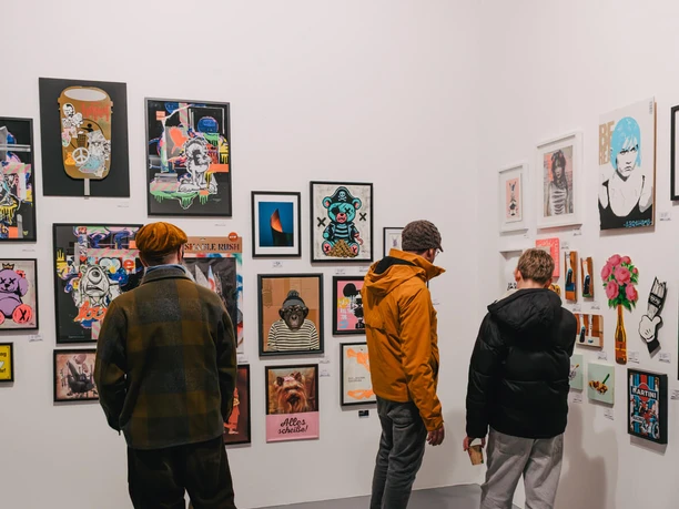 Urban Wildcard Art at Studio 40 Cologne Three people look at colorful illustrations and pictures on the white walls of a gallery.