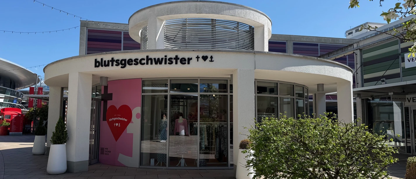 Blutsgeschwister Outlet-Shop mit runder weißer Fassade und großen Schaufenstern bei Sonnenschein.