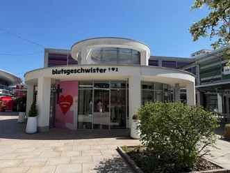 Blutsgeschwister Outlet-Shop mit runder weißer Fassade und großen Schaufenstern bei Sonnenschein.