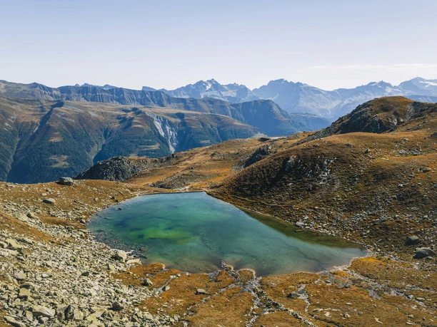 Bellwald Sommer Landschaft Web LowRes-71.jpg Teil der 7-Seen Wanderung. Klarer Bergsee inmitten einer herbstlich gefärbten, steinigen Hochlandschaft vor einer Kette markanter Alpenberge unter blauem Himmel.