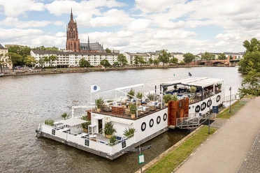 Freigut Bootshaus - Außenansicht Schiff mit Terrasse am Main, Frankfurter Dom im Hintergrund
