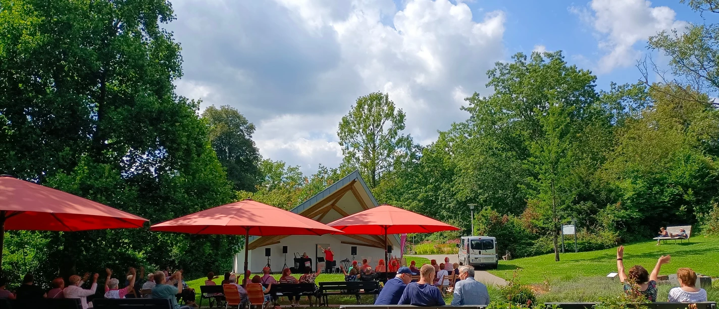 Kurkonzert im Kurpark Eckenhagen Open-Air-Konzert im Park mit Bühne, roten Sonnenschirmen und Publikum auf Bänken