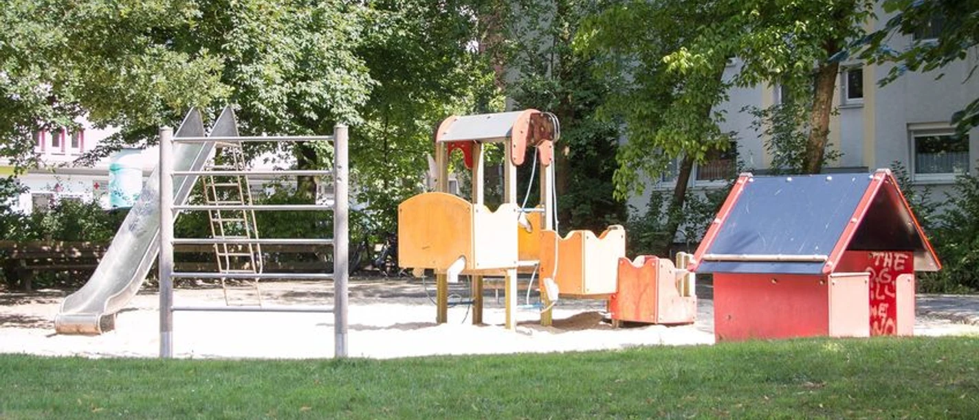 Kinderspielplatz mit Rutsche, Klettergerüst und Spielhaus auf einem Sandbereich umgeben von Bäumen