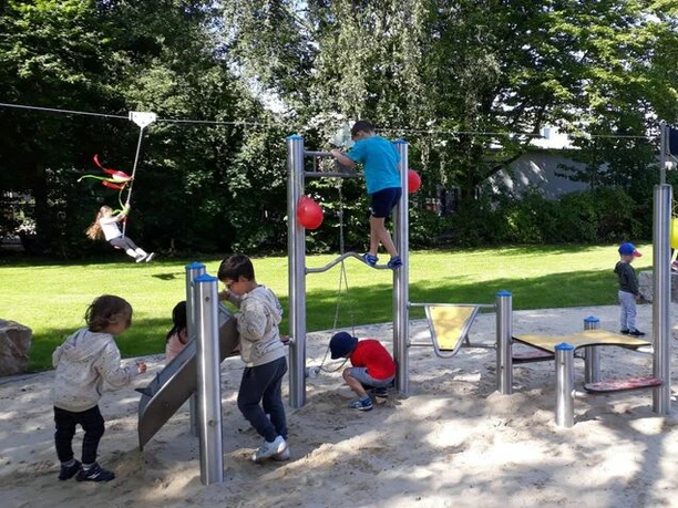 Einweihung bei strahlendem Sonnenwetter (1)_WEB.jpg Kinder spielen im Sandbereich auf dem Spielplatz und ein weitere Kind saust im Hintergrund mit der Seilbahn über den Spielplatz