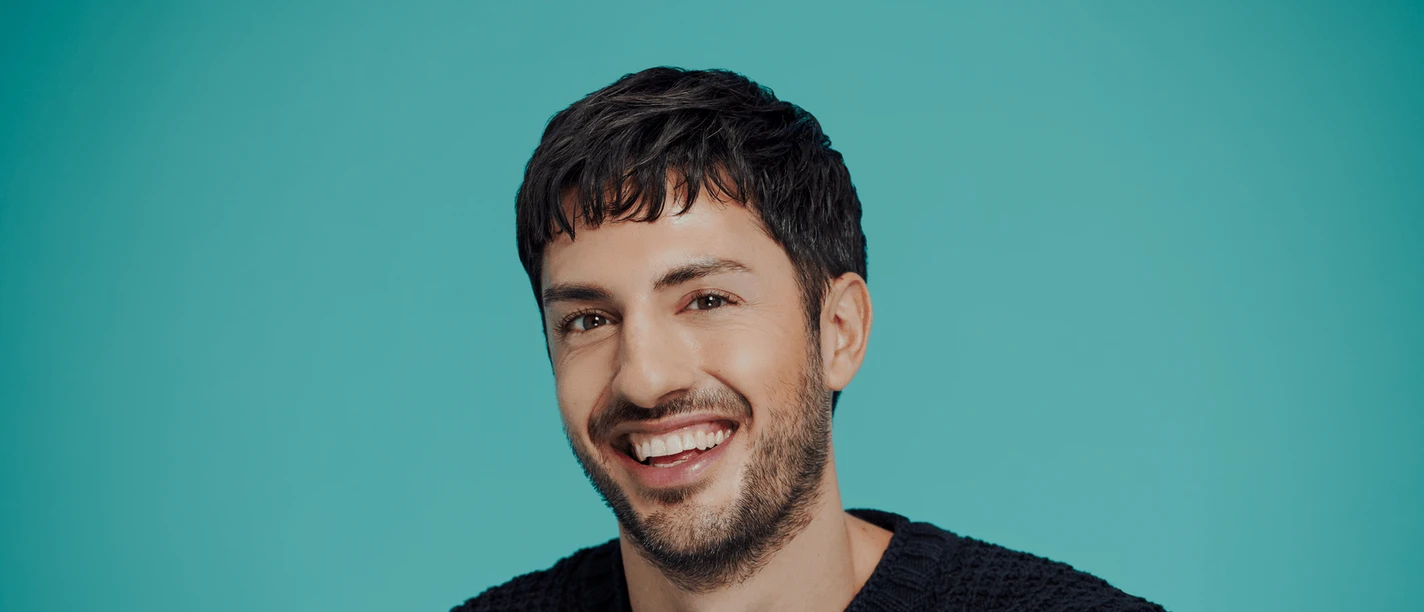 Alyosha Muttardi Smiling person with dark hair in front of a turquoise background in close-up
