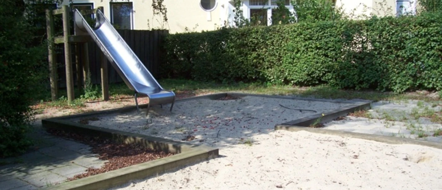 Sandspielplatz mit Rutsche, umgeben von Hecke und Bäumen