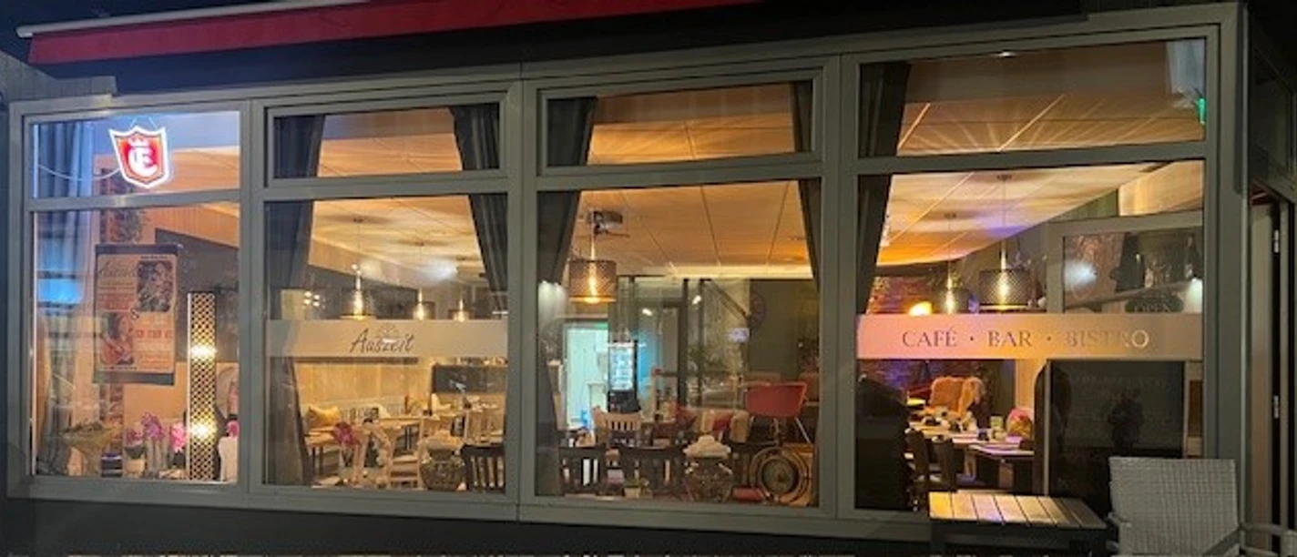 cafe-auszeit.jpg Beleuchtetes Café Auszeit mit Sitzplätzen im Innenraum und Terrasse am Abend