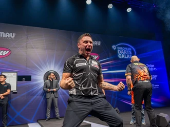 Ein Dartspieler jubelt lautstark auf der Bühne einer hell ausgeleuchteten Arena.