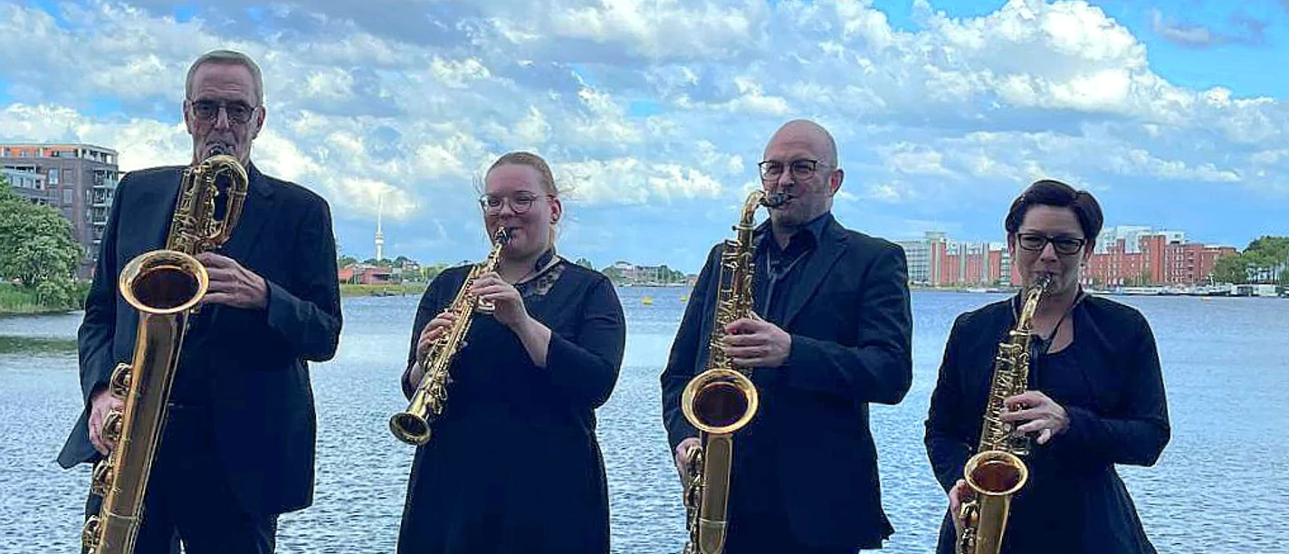 Das Quartett Vier Personen in schwarzer Kleidung spielen Saxophon vor einem Gewässer unter bewölktem Himmel.