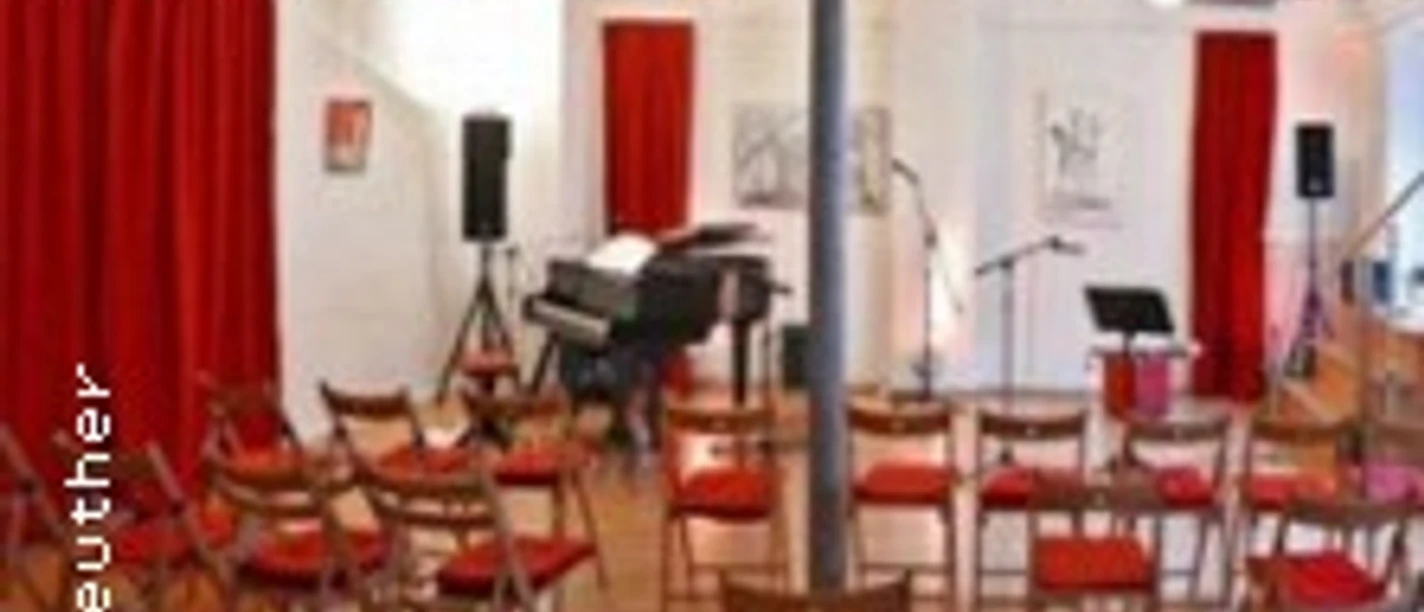 Kleiner Saal mit roten Vorhängen, Holzstühlen und einem Klavier für Aufführungen.