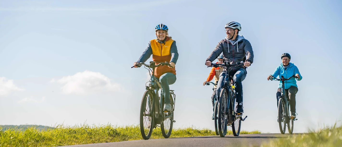 Radtour im Bergischen Vier Personen radeln entspannt auf einem Weg durch offene Wiesen unter blauem Himmel.