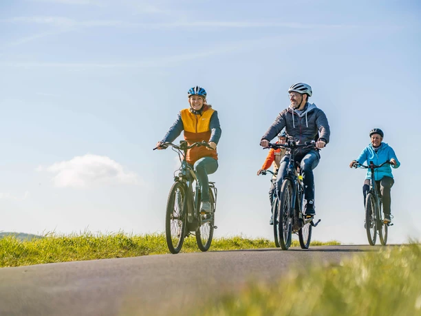 Radtour im Bergischen Vier Personen radeln entspannt auf einem Weg durch offene Wiesen unter blauem Himmel.