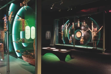 Blick in die Ausstellung - GRASSI Museum für Völkerkunde Blick in einen Ausstellungsraum des GRASSI Museums für Völkerkunde mit einer Sitzmöglichkeit und verschiedenen Vitrinen, in denen Ausstellungsstücke zu Klanggeschichten von den Andaman und Nicobar Islands präsentiert werden.
