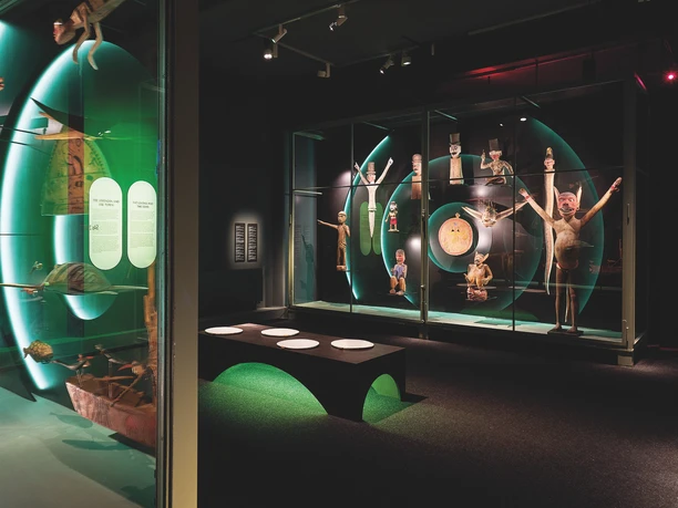 View into an exhibition room of the GRASSI Museum of Ethnology with a seating area and various showcases in which exhibits on sound stories from the Andaman and Nicobar Islands are presented.