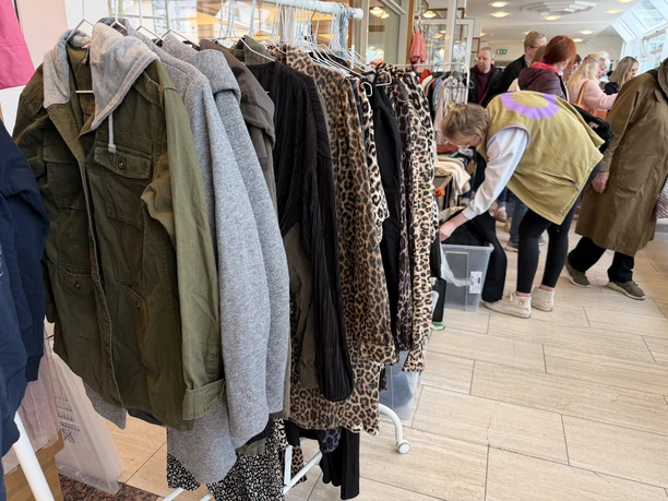 Frauenflohmarkt_1.jpeg Im Vordergrund steht ein Kleiderständer und im Hintergrund schaut eine Dame in einer Kiste mit Kleidung