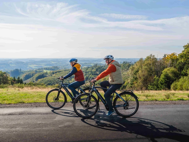 Radtour durchs Bergische Zwei Radfahrende auf einer Straße vor weitem Hügelland unter blauem Himmel