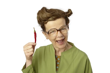 PF_Frieda Braun Sprechpause Chili nah_Foto Britta Schüssling.jpg Lachende Frieda Braun mit Retro-Brille hält amüsiert eine rote Chilischote hoch.