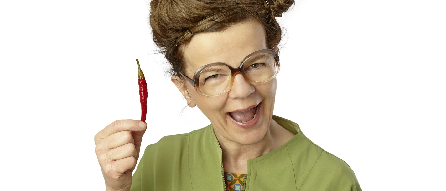 PF_Frieda Braun Sprechpause Chili nah_Foto Britta Schüssling.jpg Lachende Frieda Braun mit Retro-Brille hält amüsiert eine rote Chilischote hoch.