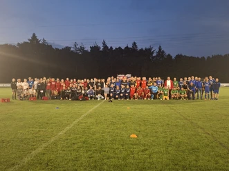 20250718_222232.jpg Fußballmannschaften posieren abends auf dem beleuchteten Rasenplatz für ein Gruppenfoto.