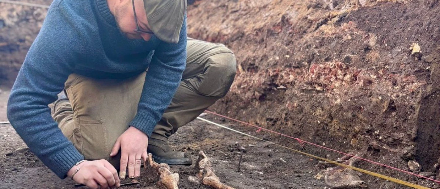 In einer Grube in der Erde arbeitet ein Archäologe an der Ausgrabung eines Skeletts. Oberhalb der Grube steht das Theater Aachen mit seiner historischen, weißen Fassade.