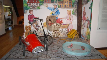 Ausstellung_Kinderwelt_Salder_HD_16zu9.jpg Rotes Tretauto mit Teddy, Puppenwagen und historisches Spielzeug vor bemalter Wand.
