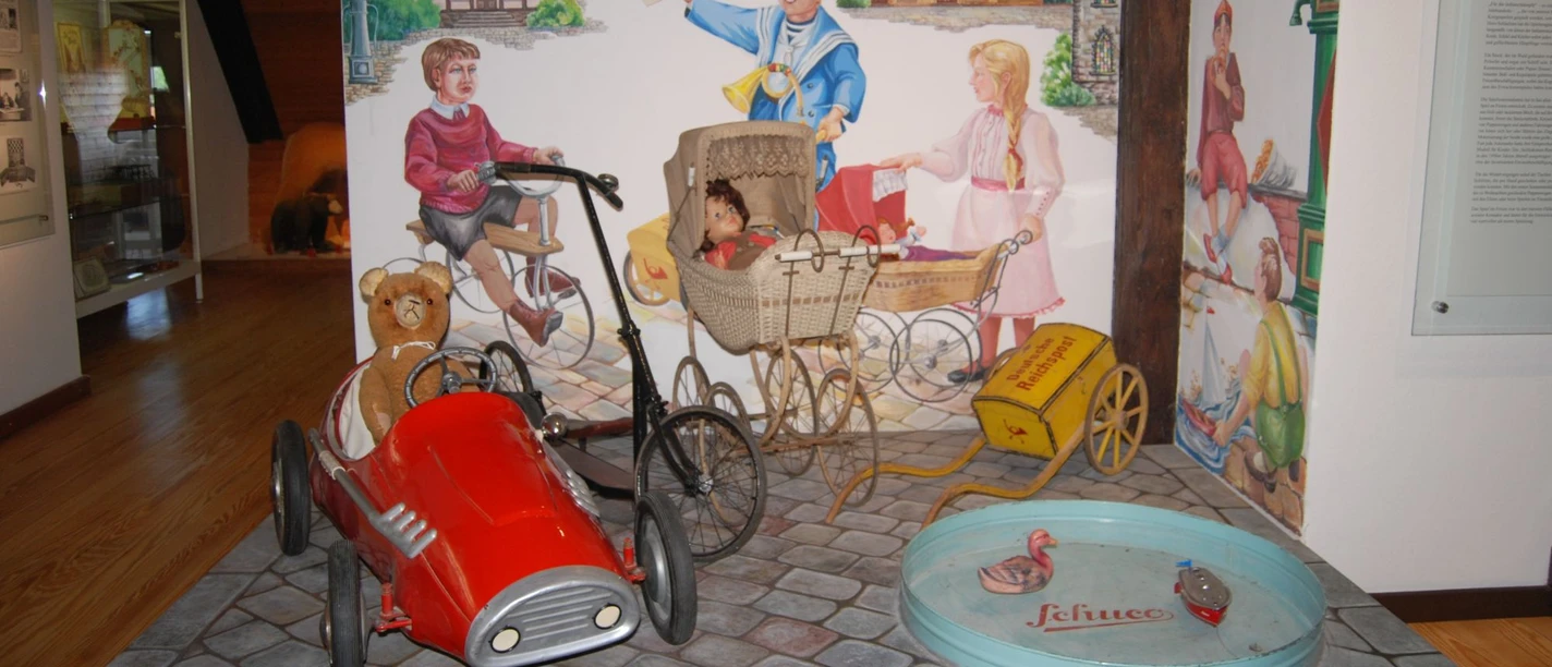Ausstellung_Kinderwelt_Salder_HD_16zu9.jpg Rotes Tretauto mit Teddy, Puppenwagen und historisches Spielzeug vor bemalter Wand.
