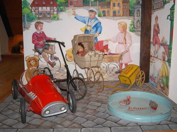 Ausstellung_Kinderwelt_Salder_HD_16zu9.jpg Rotes Tretauto mit Teddy, Puppenwagen und historisches Spielzeug vor bemalter Wand.
