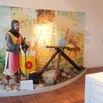 Ausstellung_Mittelalter_Salder_HD_16zu9.jpg Ritterfigur in Rüstung mit Schild und Speer vor gemalter Burgkulisse in einer Ausstellung.