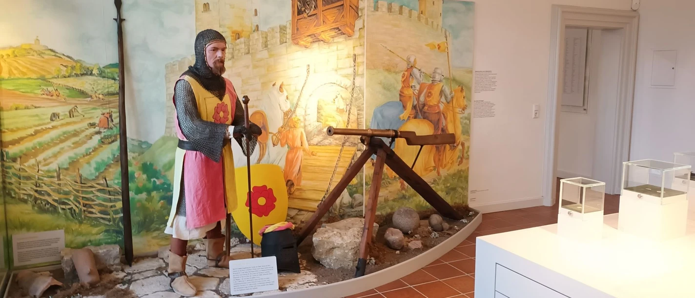 Ausstellung_Mittelalter_Salder_HD_16zu9.jpg Ritterfigur in Rüstung mit Schild und Speer vor gemalter Burgkulisse in einer Ausstellung.