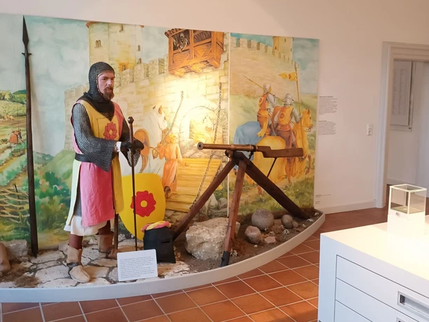 Ausstellung_Mittelalter_Salder_HD_16zu9.jpg Ritterfigur in Rüstung mit Schild und Speer vor gemalter Burgkulisse in einer Ausstellung.