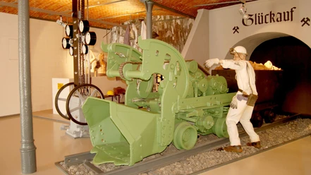 Ausstellung_Bergbau_Salder_HD_16zu9.jpg Bergmann-Figur neben grüner Bergbaumaschine vor einem Stollen mit Glückauf-Schriftzug.