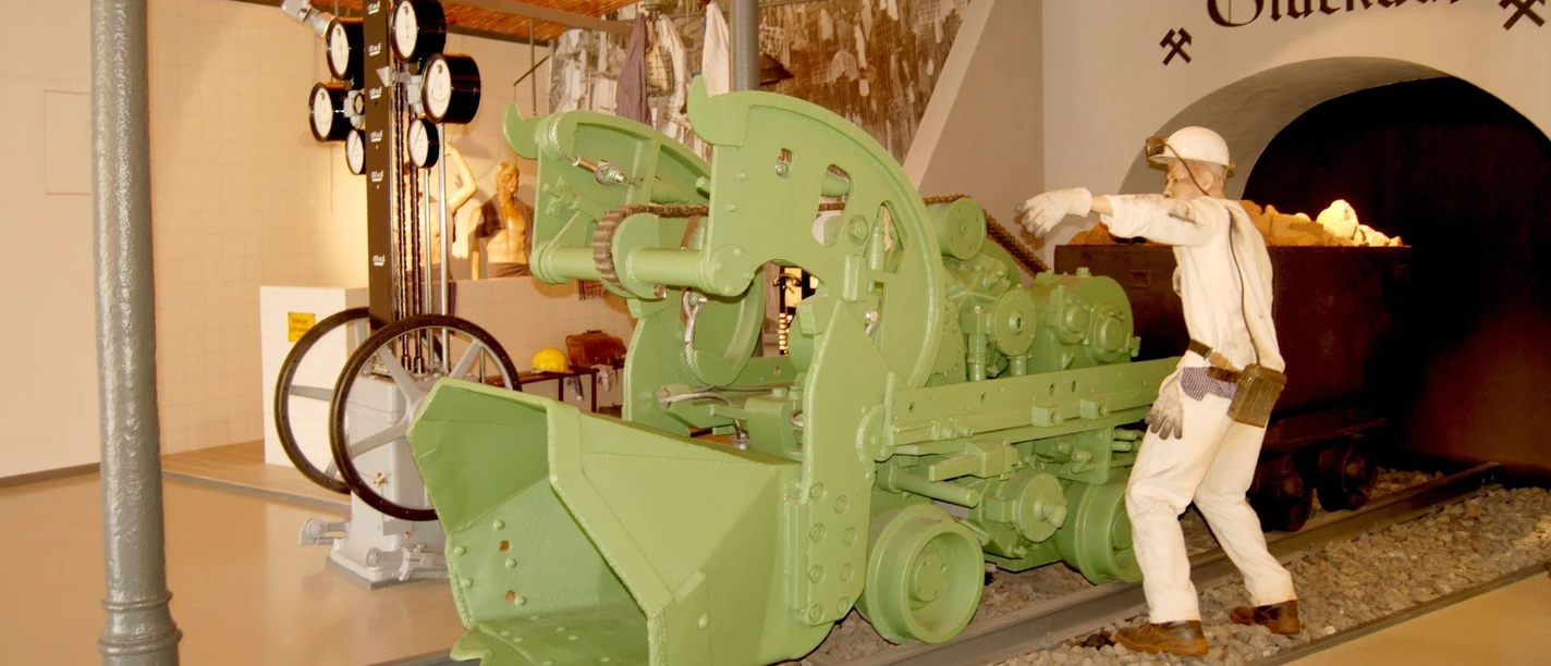 Ausstellung_Bergbau_Salder_HD_16zu9.jpg Bergmann-Figur neben grüner Bergbaumaschine vor einem Stollen mit Glückauf-Schriftzug.