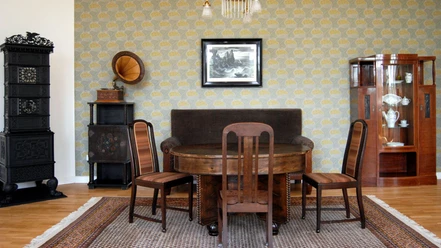 Ausstellung_Republik_Salder_HD_16zu9.jpg Historisches Zimmer mit Holzmöbeln, Grammophon und schwarzem Ofen vor gemusterter Tapete.