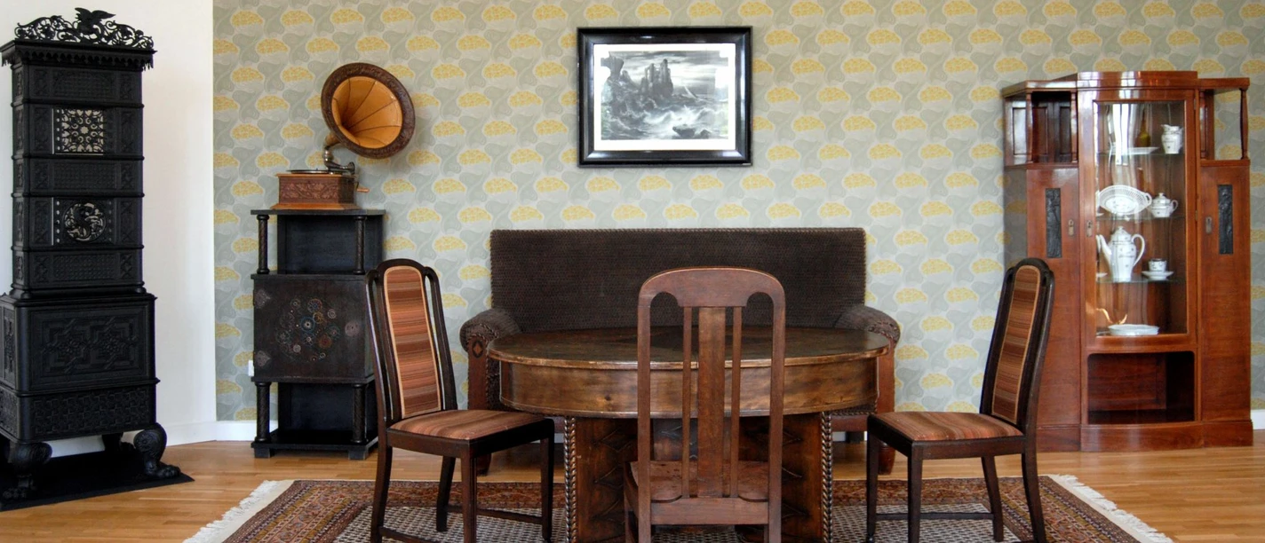 Ausstellung_Republik_Salder_HD_16zu9.jpg Historisches Zimmer mit Holzmöbeln, Grammophon und schwarzem Ofen vor gemusterter Tapete.