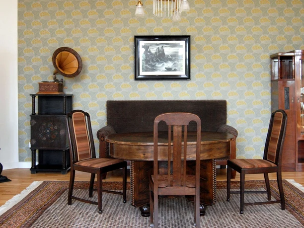Ausstellung_Republik_Salder_HD_16zu9.jpg Historisches Zimmer mit Holzmöbeln, Grammophon und schwarzem Ofen vor gemusterter Tapete.