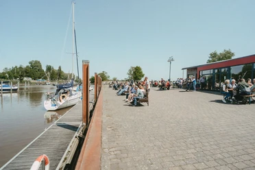 Kulinarische Meile Lebhaftes Hafenfest: Besucher speisen an der sonnigen Promenade neben ankernden Segelbooten.