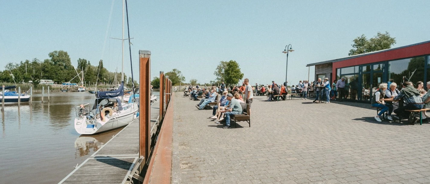 Kulinarische Meile Lebhaftes Hafenfest: Besucher speisen an der sonnigen Promenade neben ankernden Segelbooten.