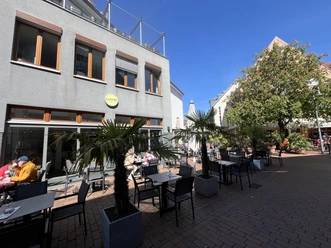La Perla Außenterrasse des Eiscafés mit Tischen, Palmen und Gästen vor modernem Gebäude