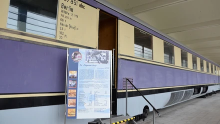 ALSTOM Werksmuseum_HD_16zu9.jpg Lila-cremefarbener historischer Zugwaggon mit geöffneter Tür und einer Informationstafel.